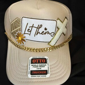 Puede incluir: Gorra de camionero color crema con parte trasera de malla y visera curva. La parte delantera presenta un parche con las palabras "Let thems" en escritura marrón, una cruz dorada y una cadena dorada. Una etiqueta dice "OTTO TRUCKER".