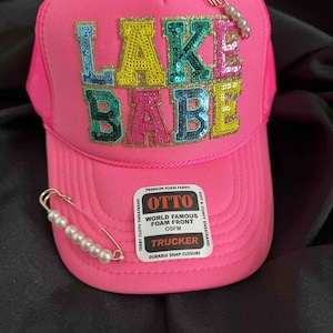 May include: A bright pink trucker hat with the words "LAKE BABE" in sequined letters. The hat has a mesh back and a curved brim. Two decorative pearl-studded pins are attached to the hat. The hat has a "OTTO" label.