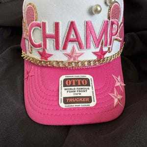 May include: A pink and white trucker hat with the word "CHAMP" in pink embroidery. The hat features gold chain trim, star accents, and tennis racket designs. The hat has an "OTTO" label.