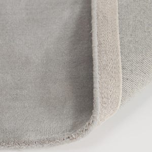 May include: A close-up of a gray rug with a soft, textured surface. The rug's edge reveals a beige herringbone pattern border. The rug is rolled up, showing the underside with a woven texture.