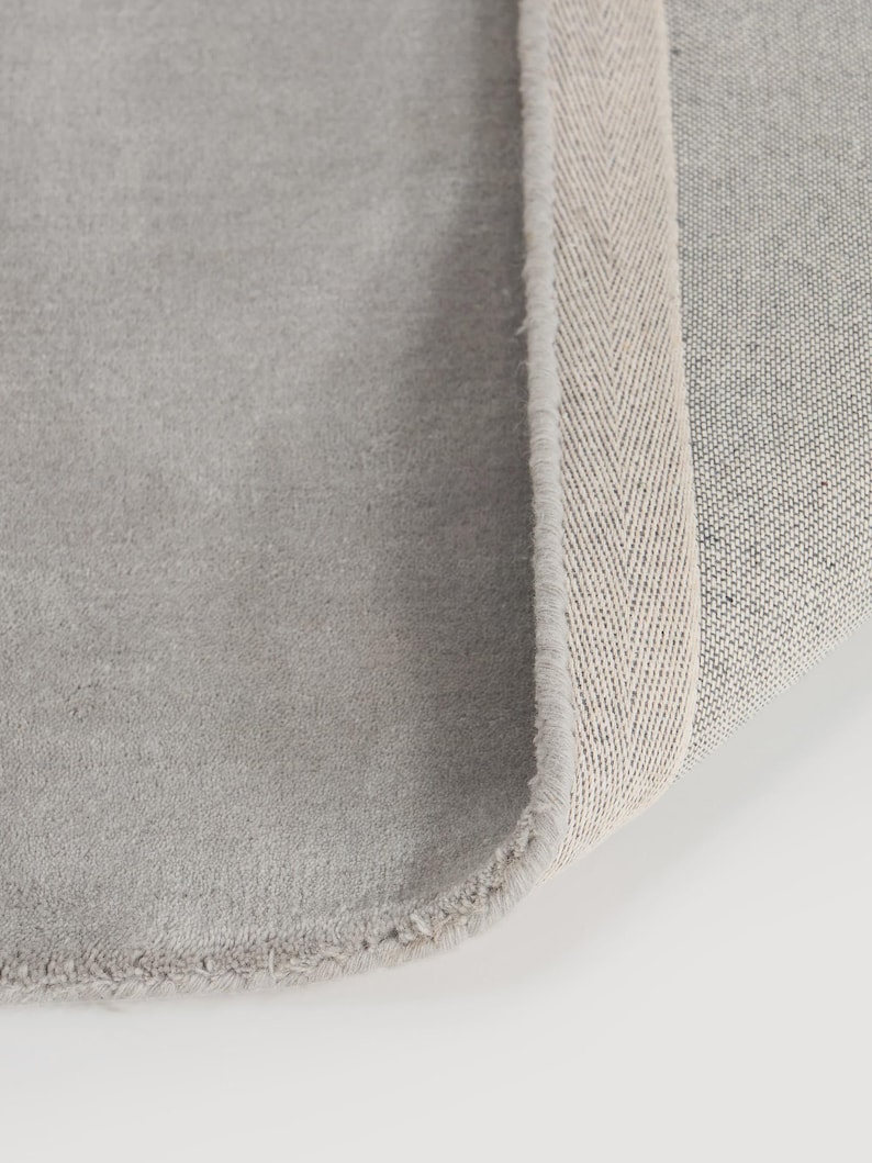 May include: A close-up of a gray rug with a soft, textured surface. The rug's edge reveals a beige herringbone pattern border. The rug is rolled up, showing the underside with a woven texture.