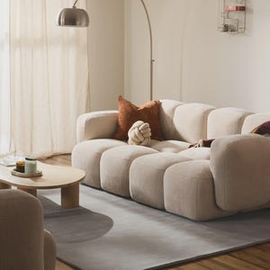 May include: A modern living room featuring a beige sectional sofa with a gray rug and wooden floor. Decorative pillows in brown and white tones rest on the sofa. A silver arc lamp and a small wooden coffee table complete the scene.