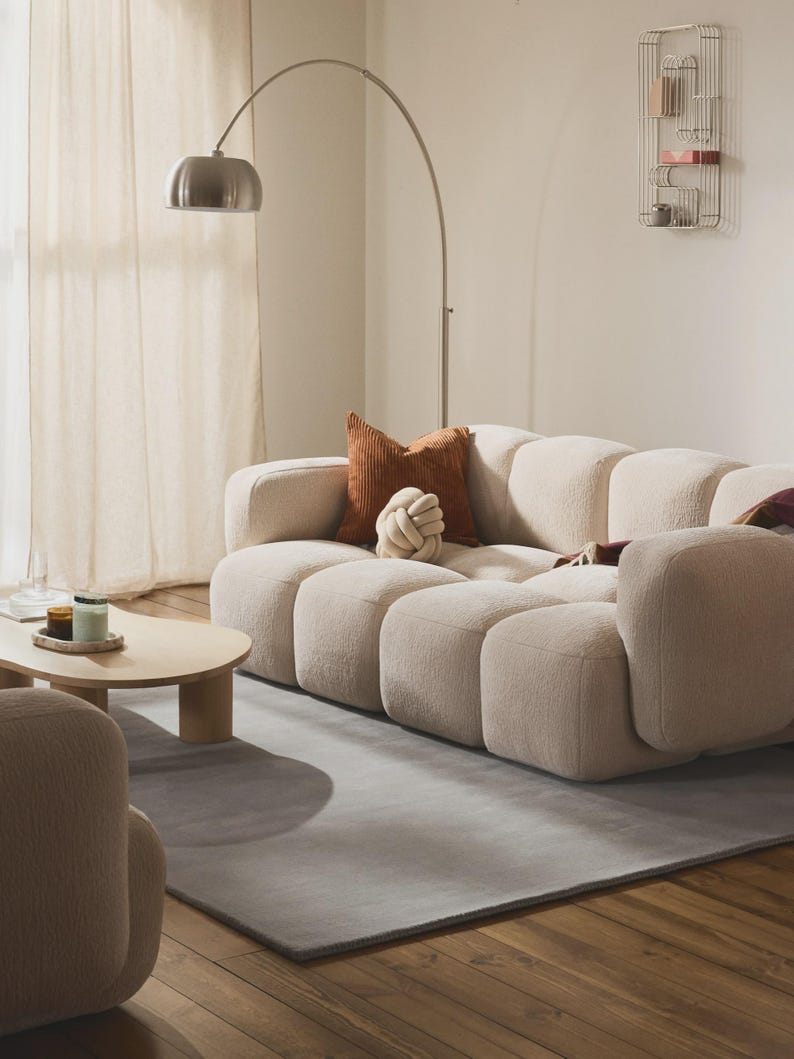 May include: A modern living room featuring a beige sectional sofa with a gray rug and wooden floor. Decorative pillows in brown and white tones rest on the sofa. A silver arc lamp and a small wooden coffee table complete the scene.