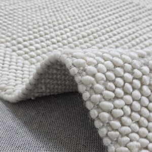 Può includere: Primo piano di un tappeto strutturato color crema con un design a pelo riccio. Il bordo del tappeto è ripiegato, rivelando il lato inferiore e la costruzione tessuta. Il tappeto è realizzato in un materiale morbido e soffice.
