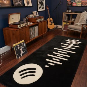 Op de afbeelding: Zwart rechthoekig tapijt met een wit Spotify-logo en golfpatroon. Het tapijt ligt op een donkere houten vloer in een kamer met een platenspeler, vinylplaten en een gitaar.