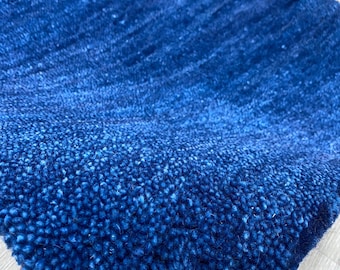 May include: A close-up of a section of a blue rug with a textured surface. The rug has a gradient effect, transitioning from a darker shade to a lighter shade of blue. The rug is held by a hand.