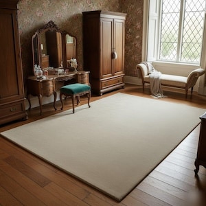 May include: A large, rectangular beige rug dominates the room's center, set on a hardwood floor. The room features antique wooden furniture, including a vanity, wardrobe, and a chaise lounge near a window. The walls are adorned with floral wallpaper.