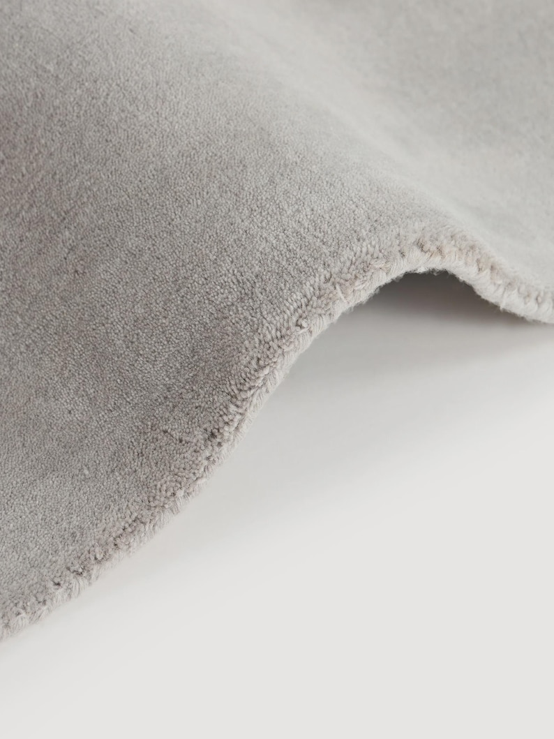 May include: Close-up of a section of a light gray rug with a soft, textured surface. The edge of the rug is visible, showing a neatly finished border. The rug is folded over, revealing its thickness and construction.