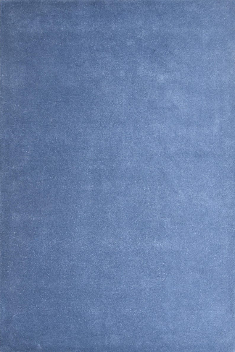 May include: A solid, rectangular, blue rug with a soft, velvety texture. The rug has a muted, dusty blue color, and the surface appears to have subtle variations in tone, creating a sense of depth. The rug is suitable for home decor.