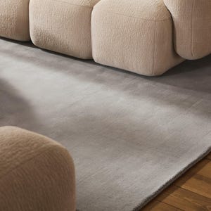 May include: A modern living room scene featuring a light gray rug and a beige modular sofa. The rug has a subtle texture and a dark gray border. The sofa has a rounded, textured design, creating a cozy and inviting atmosphere.