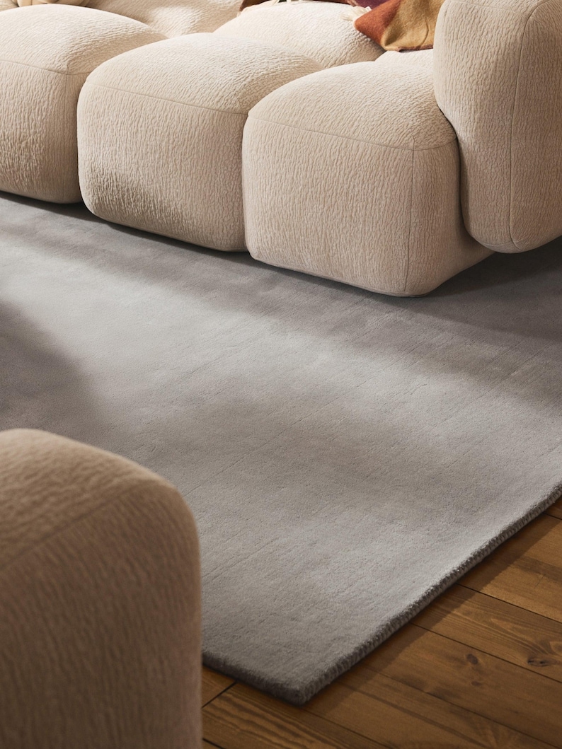 May include: A modern living room scene featuring a light gray rug and a beige modular sofa. The rug has a subtle texture and a dark gray border. The sofa has a rounded, textured design, creating a cozy and inviting atmosphere.