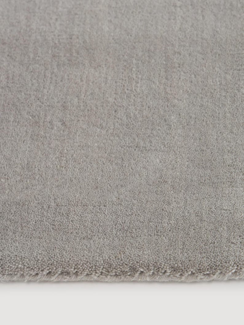 May include: A close-up of a light gray rug with a soft, textured surface. The rug has a subtle, uniform color and a slightly blurred edge, suggesting a plush, comfortable texture. The neutral tone makes it suitable for various interior design styles.