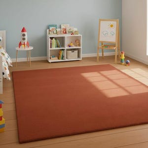 May include: A large, rectangular, rust-colored rug dominates the floor of a child's bedroom. The room features a white bed with a colorful dinosaur-themed comforter, a white bookshelf, and a small table with a rocket ship decoration. A wooden easel and toys are also present.