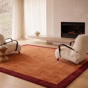 May include: An orange area rug with a deep red border dominates the room. Two white, fluffy armchairs with dark wooden frames sit on the rug. A wooden side table and a fireplace complete the cozy, modern living room setting.