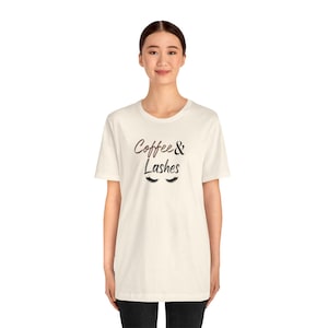 May include: Cream-colored t-shirt with the text "Coffee & Lashes" in a handwritten style, with a pair of stylized eyelashes below the text. The shirt is a casual, everyday wear item.