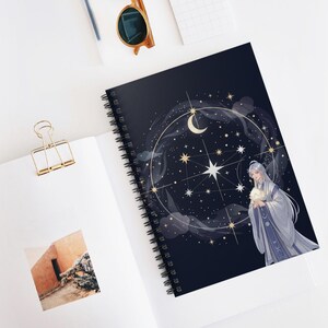 Könnte beinhalten: Spiralgebundenes Notizbuch mit dunkelblauem Einband mit einem Himmelsdesign. Das Cover-Artwork zeigt einen Halbmond, Sterne und eine Illustration einer Person in weißen Roben. Das Notizbuch ist auf einer weißen Oberfläche geöffnet.
