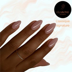 Könnte beinhalten: Eine Hand mit mandelförmigen Nägeln, die in einem hellrosa Farbton lackiert sind. Die Nägel haben ein glänzendes Finish. Ein goldener Ring mit kleinen Steinen befindet sich am Ringfinger. Der Text "Classique signature" und das Logo Clarone Studio sind enthalten.