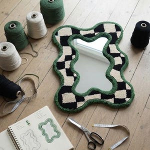Checkerboard tufted mirror displayed on a wooden floor with yarn cones, tufting tools, scissors, and a design sketch.