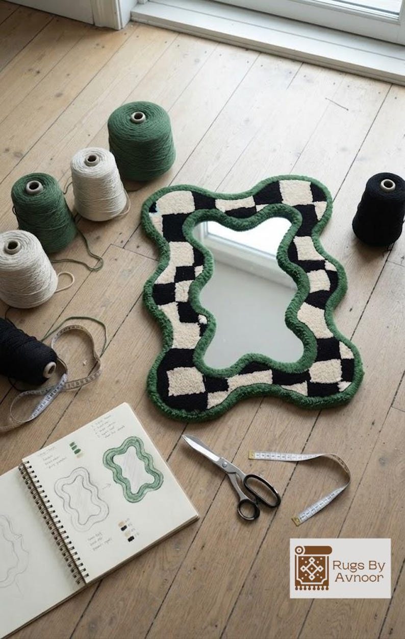 Checkerboard tufted mirror displayed on a wooden floor with yarn cones, tufting tools, scissors, and a design sketch.