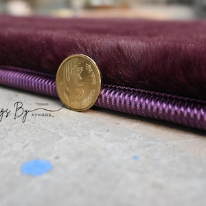 May include: Close-up of a plush, deep purple rug with a textured surface. A gold coin rests on the edge, providing a size reference. The rug has a decorative purple border. The text "Rugs By Avnoor" is visible.