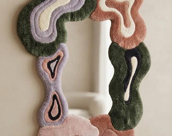 Può includere: Uno specchio da parete incorniciato da un bordo in tessuto trapuntato e morbido con forme organiche irregolari nei toni del verde oliva, lavanda, crema, terracotta e rosa tenue. L'angolo in basso a destra riporta il testo Rugs By Avnoor.