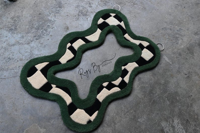 Pu&ograve; includere: Un tappeto dalla forma irregolare e ondulata con un bordo spesso verde scuro e un motivo a scacchi neri e crema. Il centro &egrave; aperto e il testo Rugs By Avnoor &egrave; stampato al centro. Due anelli in metallo argentato sono fissati nella parte superiore.