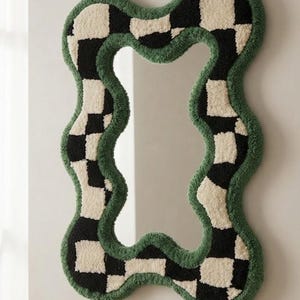 Op de afbeelding: Een wandspiegel met een onregelmatig, golvend frame bedekt met een getuft ruitpatroon van zwarte en crèmekleurige vierkanten. De buiten- en binnenranden van het frame zijn afgewerkt met een effen donkergroene getufte rand. Tekst: Rugs By Avnoor.