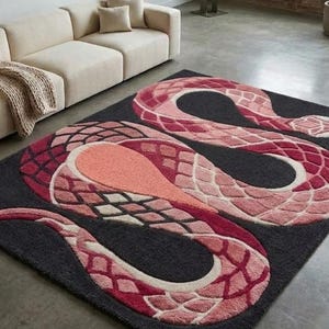 Può includere: Un tappeto rettangolare con un grande disegno di serpente stilizzato in tonalità rosa, corallo e rosso scuro su uno sfondo grigio antracite. Il serpente presenta un motivo testurizzato a scaglie su tutto il corpo. Testo: Rugs By Avnoor.