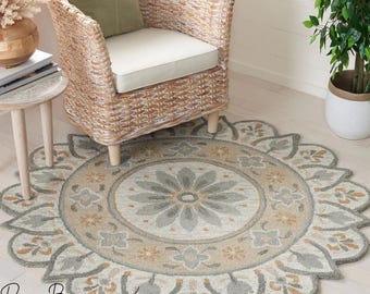 May include: A round area rug with a floral design in shades of gray, beige, and gold. The rug is placed on a light-colored wooden floor, with a wicker chair and side table in the background. The rug's design features a central flower motif.