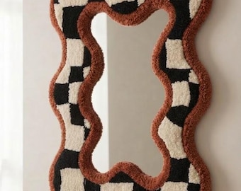 Può includere: Uno specchio da parete con una cornice ondulata e irregolare ricoperta da un motivo a scacchi trapuntato di quadrati neri e crema. Il bordo esterno della cornice è rifinito con un spesso bordo testurizzato color ruggine. Il testo Rugs By Avnoor è visibile.