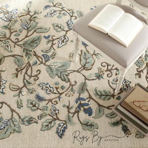 以下が含まれることがあります： 青、緑、茶色の花柄のベージュ色のラグ。葉とつるの精巧な植物デザインが特徴です。灰色の布張りの椅子と本が見えます。