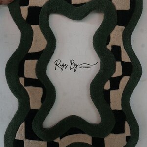Pu&ograve; includere: Un tappeto tufted dalla forma irregolare e ondulata con un motivo a scacchi neri e crema bordato da un bordo spesso verde scuro. Il centro del tappeto &egrave; vuoto e il testo Rugs By Avnoor &egrave; stampato in nero al centro.