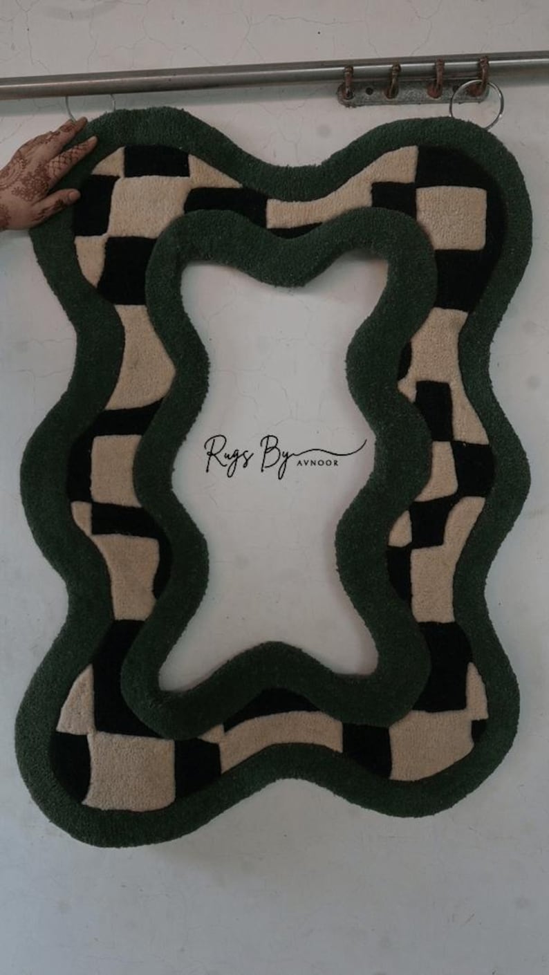 Pu&ograve; includere: Un tappeto tufted dalla forma irregolare e ondulata con un motivo a scacchi neri e crema bordato da un bordo spesso verde scuro. Il centro del tappeto &egrave; vuoto e il testo Rugs By Avnoor &egrave; stampato in nero al centro.