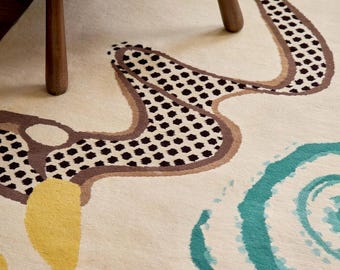 May include: A cream-coloured area rug featuring a large, winding brown and black patterned shape, a solid yellow organic form, and a circular teal swirl design. The rug has a soft, textured pile and is displayed under wooden furniture legs.