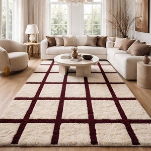Puede incluir: Una alfombra de área de color crema, afelpada, con un patrón de cuadrícula en un color burdeos intenso. La alfombra está colocada en un salón con un sofá seccional blanco, una mesa de centro redonda y suelo de madera. La habitación tiene grandes ventanas y decoración de tonos neutros.