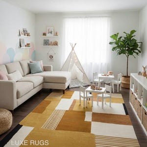 May include: A modern living room with a beige sectional sofa, white tables and chairs, and a geometric rug in shades of yellow, brown, and white. A teepee and a potted plant add to the room's decor. The rug has the text "LUXE RUGS".