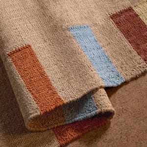 May include: Close-up of a woven rug with a beige base and colourful stripes. The stripes are in shades of orange, blue, and red. The rug has a textured appearance and is folded over, revealing the underside.