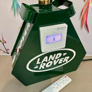 May include: A dark green, geometric-shaped speaker with the Land Rover logo in white. It has a gold-colored top and a white control panel with a blue display. A white remote control lies in front of the speaker.