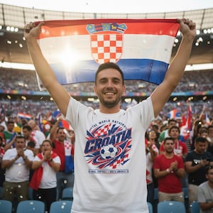 May include: A person holding a Croatian flag above their head, smiling at a stadium event. The person is wearing a white t-shirt with a Croatia soccer design. The flag has a red, white, and blue design.