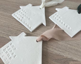 May include: Three white ceramic house-shaped ornaments with embossed text and ribbon ties. One reads "the groves home sweet home", another "the barnette", and the third "our first home 2023". The ribbons are white, beige, and green.