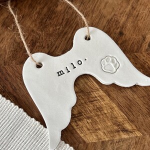 May include: White ceramic angel wings ornament with the name "milo" and a paw print. The ornament is strung with twine and is designed for pet memorial or pet loss remembrance.