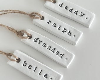 May include: Four white rectangular tags with black text and twine. The tags read "daddy", "ralph", "grandad", and "bella". Each tag has a small hole at the top for the twine. The background is white.