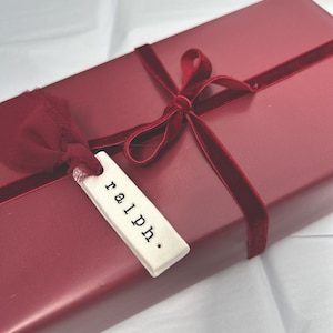 May include: A burgundy gift box, rectangular in shape, adorned with a velvet ribbon and bow. A small tag, bearing the name "ralph.", is attached. The box rests on a white surface.