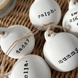 May include: A close-up shot of several white, round baubles with black text. The baubles are strung with twine and feature names such as "Ralph," "Daddy," and "Mummy." These are likely for a Christmas tree or other festive decoration.