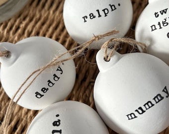 May include: Close-up of several white, round ornaments with black text. The ornaments are strung with twine and feature names like "Ralph," "Daddy," and "Mummy." The ornaments are likely for a Christmas tree or other holiday decoration.