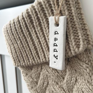May include: A beige knitted Christmas stocking with a white tag that reads "daddy." The tag is attached with twine. The stocking has a cable knit design.