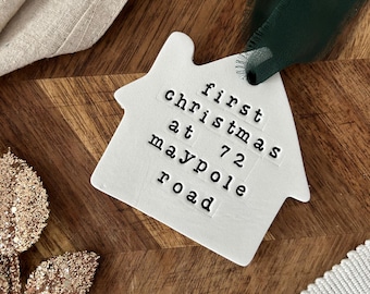 May include: A white ceramic house-shaped ornament with the text "first christmas at 72 maypole road" in black letters. A dark green ribbon is attached for hanging. The ornament is on a wooden surface with a gold leaf and white fabric.