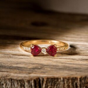 Op de afbeelding: Een gouden ring met een strikdesign, met twee hartvormige, dieprode edelstenen en een centrale, ronde, heldere edelsteen. De ringband is ook versierd met kleine, heldere edelstenen.