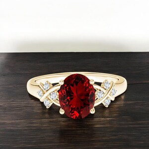 Könnte beinhalten: Ein goldener Ring mit einem ovalen, tiefroten Edelstein im Mittelpunkt. Der Ring ist mit kleinen, runden, klaren Edelsteinen besetzt und hat ein dekoratives Banddesign. Der Ring liegt auf einer dunklen Holzoberfläche.