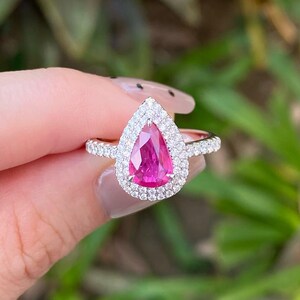 Puede incluir: Anillo con una gema rosa en forma de pera, rodeada por un halo de pequeñas piedras claras. La banda también está adornada con pequeñas piedras claras. Fondo verde borroso.